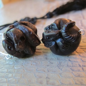 May include: Two dark brown, carved animal pendants. One pendant is a turtle, and the other is a ram. Each pendant has a small silver loop attached. A black cord and a silver wire are visible in the background.