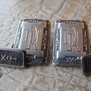 May include: A pair of silver cufflinks with a monogrammed design. The cufflinks are rectangular and have a textured border. The monogram is a stylized "JR" in a script font.