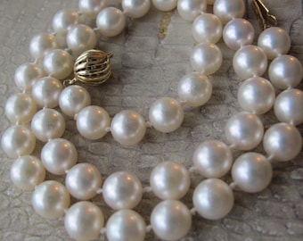 Op de afbeelding: Een dubbele rij parelketting met een gouden sluiting. De witte, ronde parels hebben een glanzende glans. De sluiting heeft een gedetailleerd, gestructureerd ontwerp. De ketting wordt gepresenteerd op een gestructureerd oppervlak.