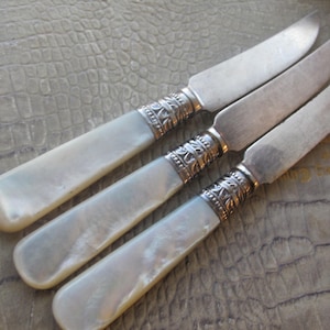May include: Three antique butter knives with mother-of-pearl handles and decorative silver collars. The stainless steel blades are arranged diagonally. These knives showcase an elegant design and fine craftsmanship.