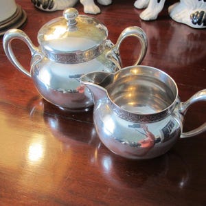 May include: A shiny silver tea set, including a teapot with a lid and a creamer. Both pieces have curved handles and decorative trim around the top. The set is displayed on a polished wooden surface.
