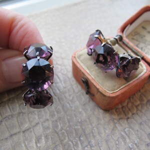 May include: A pair of vintage amethyst-colored earrings and a matching ring. The jewelry features large, faceted, purple gemstones set in a silver-toned metal. The earrings are displayed in a small, pink, velvet-lined box.