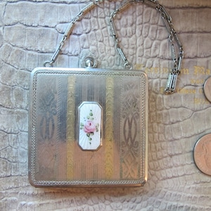 以下が含まれることがあります： チェーンが付いた、銀と金のコンパクトで、花柄のデザインが施されています。コンパクトには、白いエナメルパネルがあり、ピンクのバラが描かれています。コンパクトは、日本の100円玉くらいの大きさです。