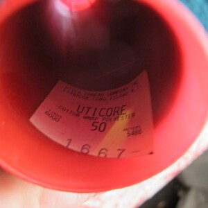 Puede incluir: Una bobina roja de hilo de poli&eacute;ster de algod&oacute;n envuelto Uticore con una etiqueta que dice "Uticore Cotton Wrap Polyester 50" y "Yards 6000 Meters 5486". La etiqueta tambi&eacute;n incluye el n&uacute;mero "1667".