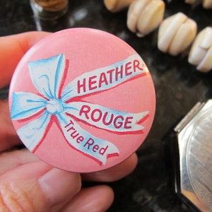 May include: A pink button featuring a blue and white bow design and the words "HEATHER ROUGE True Red" in red text. The button is held in a hand, with other objects in the background.