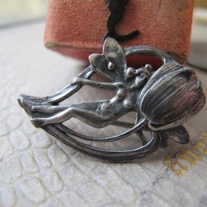May include: A silver-toned brooch featuring a fairy figure reclining within a stylized flower. The fairy has delicate wings and is positioned within a curved frame. The brooch has an antique appearance.