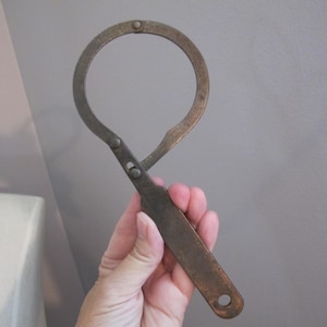 Könnte beinhalten: Ein altes Metallmesswerkzeug mit einem kreisförmigen Öffnungsmechanismus, einem beweglichen Arm und einem flachen, rechteckigen Griff. Das Werkzeug weist eine verwitterte, dunkel bronzefarbene Patina auf, die deutliche Gebrauchsspuren zeigt.