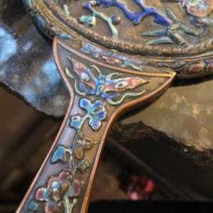 May include: Antique hand mirror with a detailed floral and butterfly design. The handle and frame are bronze-colored, with colorful enamel accents in blue, pink, and green. The mirror's surface is not visible.