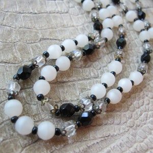 May include: A long necklace made of white and black beads. The white beads are opaque and round, while the black beads are faceted and have a shiny finish. The necklace is strung on a silver chain.