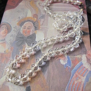 May include: A clear, faceted crystal bead necklace featuring a series of transparent, multi-sided beads strung together. The necklace includes a small, dark metal circular clasp at the end, providing a secure closure for the strand of reflective beads.