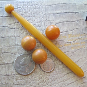 Op de afbeelding: Vier ronde amberkleurige kralen en een lange, dunne amberkleurige stok. De stok heeft een ronde amberkleurige kraal aan het ene uiteinde. De kralen en de stok liggen op een lichtbruin, getextureerd oppervlak.