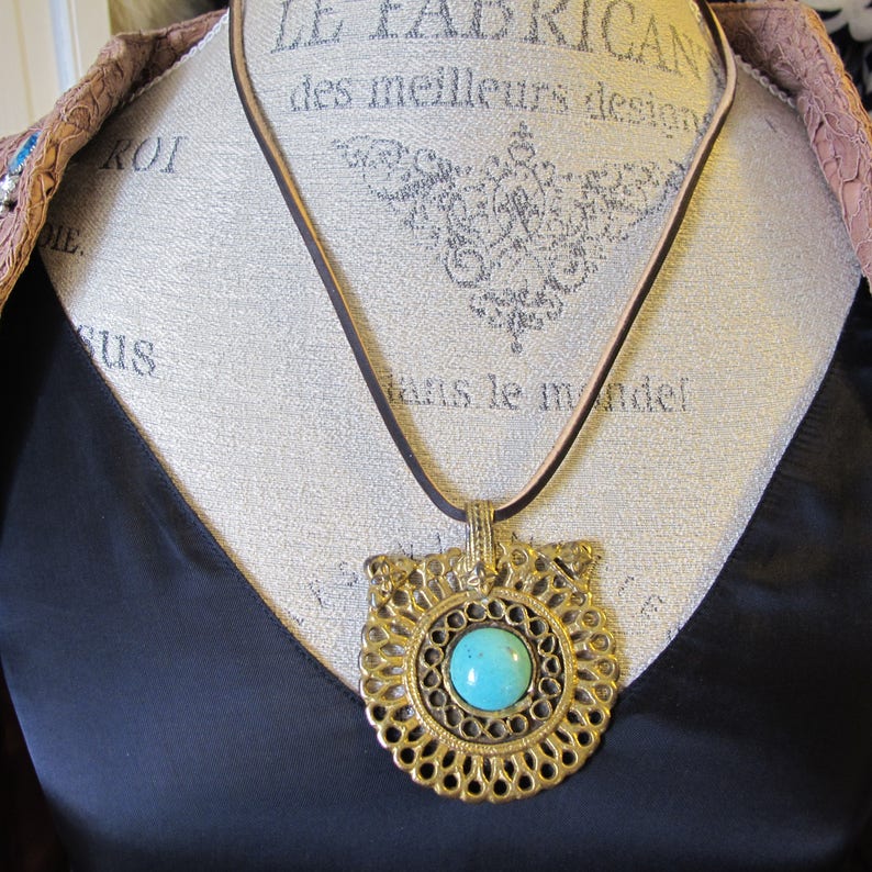 Op de afbeelding: Een turquoise edelsteen hangerketting met een gouden filigraan ontwerp. De ketting is aan een bruin leren koord.