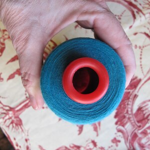 Puede incluir: Una bola de hilo azul turquesa con una bobina de pl&aacute;stico roja en el centro. El hilo est&aacute; siendo sostenido por una mano.