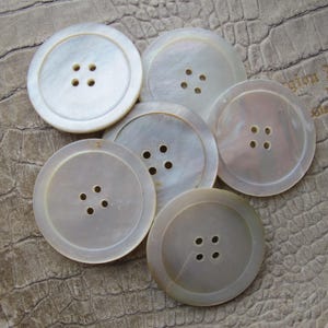 May include: Six white mother-of-pearl buttons, each with four holes. The buttons are round and have a raised edge.