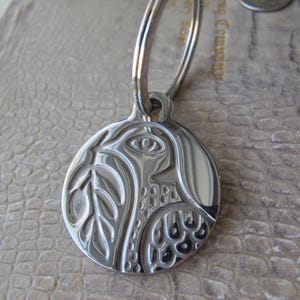 Op de afbeelding: Zilverkleurige hanger met een gedetailleerd gegraveerd ontwerp van een vogel en bladeren. De ronde hanger heeft een lus voor een ketting of sleutelhanger. Het ontwerp bevat een oog, bladeren en gedetailleerde patronen.