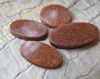 May include: Four oval-shaped sandstone cabochons in shades of brown and orange with a glittery finish. The cabochons are arranged on a textured surface.