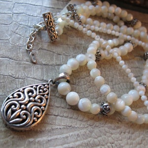 Puede incluir: Un collar de perlas blancas con un colgante de plata con un diseño de espiral. El collar tiene un cierre de plata y está formado por dos hileras de perlas.