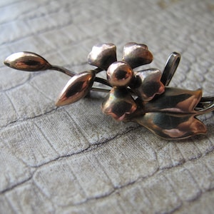 May include: A silver and gold toned flower brooch with a single flower and leaves. The flower has five petals and a center with a raised circle. The leaves are long and narrow.