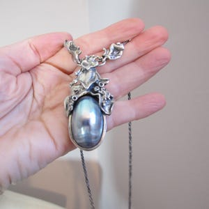 Peut inclure: Un collier argenté orné d'un grand pendentif perle ovale irisé. Le pendentif est serti dans un cadre décoratif d'inspiration florale. La chaîne est de couleur argent foncé.