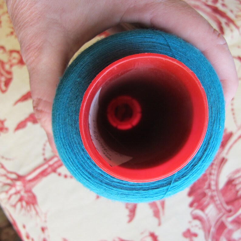 Puede incluir: Una bobina de hilo azul turquesa con un centro de pl&aacute;stico rojo. El hilo est&aacute; enrollado firmemente alrededor de la bobina.