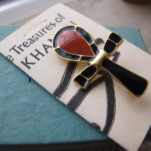 May include: A gold-toned ankh pin with black and red enamel detailing. The ankh is set on a cream-colored tag with the text "The Treasures of KHA" printed on it. The pin is resting on a blue-green surface.