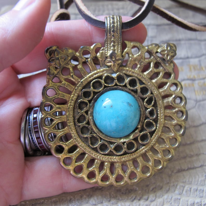 Op de afbeelding: Een gouden ketting met een hanger met een turquoise steen in een cirkelvormig ontwerp. De hanger is versierd en heeft een gedetailleerd filigraanpatroon. De ketting is van leer.