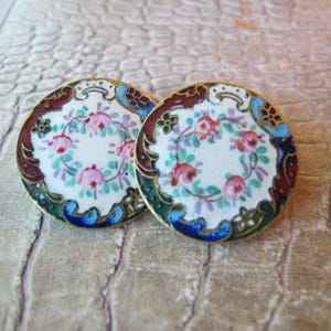 May include: A pair of round, decorative buttons with a white background and a floral design in pink, green, blue, and purple. The buttons have a gold rim and a blue and green border.