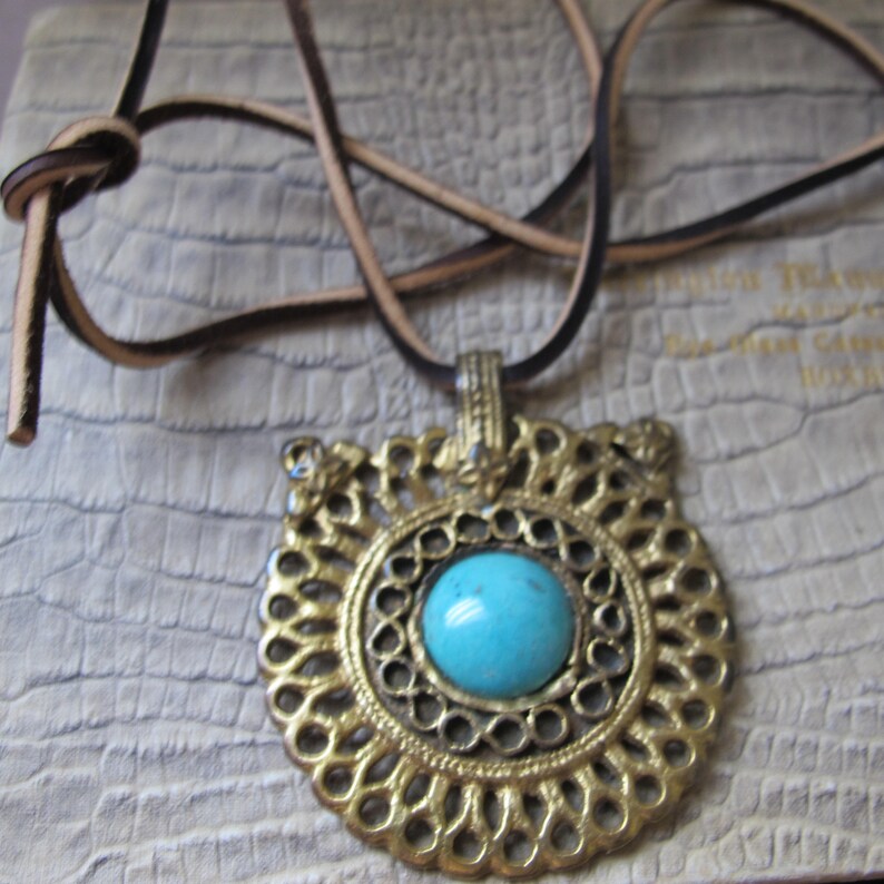 Op de afbeelding: Een turquoise edelsteen hanger ketting met een gouden filigraan ontwerp. De ketting is aan een bruin leren koord.