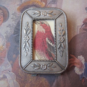 May include: A rectangular silver-toned metal picture frame with a textured surface and beaded edges. The frame features embossed floral and leaf motifs on all four sides and contains a small illustration of a red bird with white and black wings.