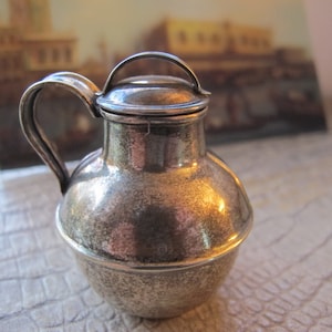 Op de afbeelding: Een zilveren kan met een deksel en een handvat. De kan heeft een getextureerd oppervlak en een rond lichaam.