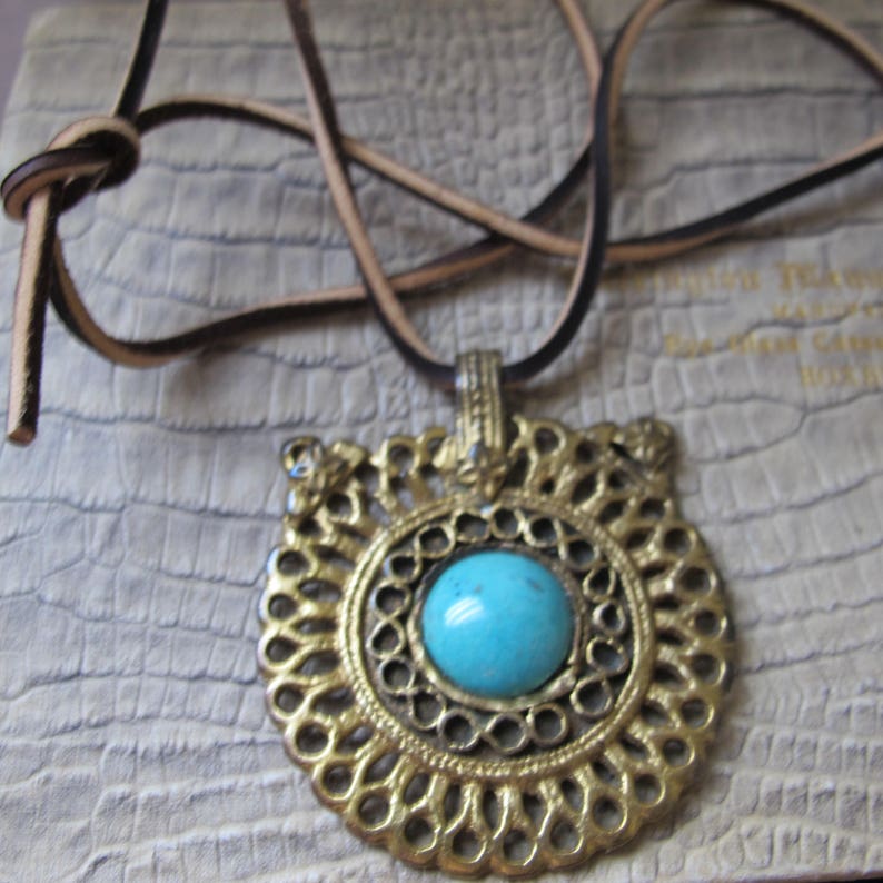 Op de afbeelding: Een turquoise edelsteen hangerketting met een gouden filigraanontwerp. De ketting is aan een bruin leren koord.