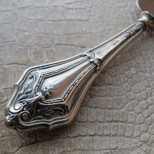 Op de afbeelding: Een zilveren handvat met een sierlijk ontwerp, met ingewikkelde bloemmotieven en een gedetailleerd, gestructureerd oppervlak. Het handvat is waarschijnlijk voor een serveerbestek of een decoratief stuk.