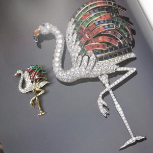 Op de afbeelding: Twee flamingo-vormige broches met heldere kristallen en veelkleurige baguette-geslepen stenen. De grotere broche is zilverkleurig met heldere kristallen, terwijl de kleinere broche een goudkleurig lichaam en poten heeft.