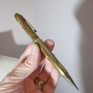 May include: A vintage gold-toned pen with a textured, ridged design. The pen features a clip and a pointed tip. The pen is held in a hand, showcasing its size and design. The pen's body has a light and dark gold color.