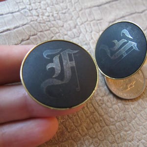 May include: A pair of black cufflinks with gold trim. Each cufflink features a silver-colored, stylized letter 'F' in a gothic font.