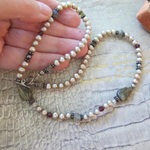 May include: A necklace featuring a strand of round, cream-colored pearls interspersed with small, dark red beads, metallic spacers, and two carved, leaf-shaped grey stones. The piece is secured with a silver-toned toggle clasp at the ends.