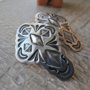 May include: A pair of silver-coloured metal conchos with detailed engraved designs. Each concho has a central diamond shape and surrounding floral and geometric patterns. They are shown on a textured, neutral-toned surface.