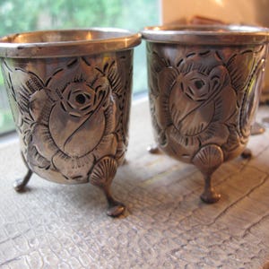 May include: Two silver-toned metal cups featuring an embossed rose pattern on the exterior. Each cup has a smooth, rounded rim and is supported by three small, shell-shaped feet at the base. The metal surface shows a polished, reflective finish.