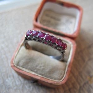 May include: A silver ring with a row of six vibrant red gemstones, likely rubies, set in a delicate, openwork design. The ring is displayed inside a vintage pink jewelry box with a soft, velvet interior.