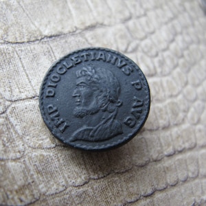 May include: A black metal coin with a profile of a man wearing a helmet and the text "IMP DIOCLETIANS P AVG" around the edge.