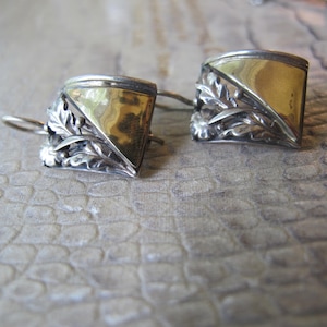 May include: A pair of silver and gold-toned earrings. Each earring features a square design with a gold-colored center and silver filigree detailing. The silver elements include floral and leaf patterns. The earrings have a hook closure.