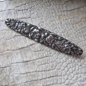 May include: A silver-toned, ornate, floral-shaped brooch with a detailed design of roses and leaves.