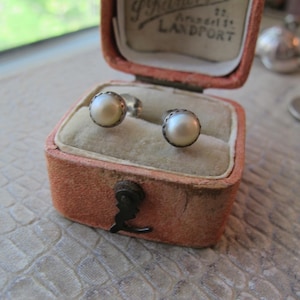 Puede incluir: Pendientes de perlas en una caja de joyería antigua de color melocotón. Los pendientes tienen un engaste plateado alrededor de las perlas blancas. La caja está forrada con terciopelo beige claro y tiene un cierre metálico.