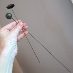 May include: Two vintage hat pins with black metal tops and round green glass heads. The pins are held in a hand against a light gray background.