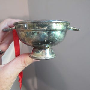 May include: A miniature silver-toned metal colander ornament featuring a rounded bowl, a pedestal base, and two small side handles. A thin red ribbon is attached to one handle for hanging, and the bowl has small drainage holes visible.