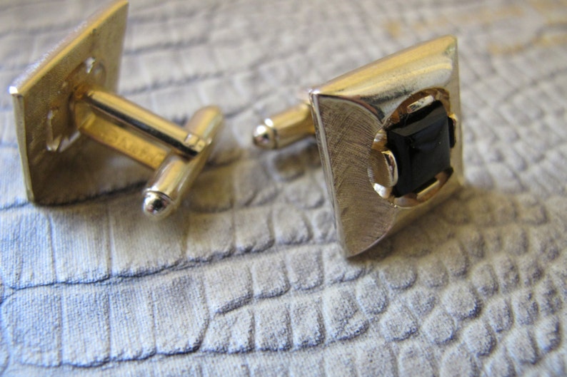 K&ouml;nnte beinhalten: Ein Paar goldfarbene Manschettenkn&ouml;pfe mit quadratischem Design. Jeder Manschettenknopf hat eine geb&uuml;rstete Metalloberfl&auml;che und einen schwarzen, quadratisch geschliffenen Stein in einem goldenen Rahmen. Die Manschettenkn&ouml;pfe werden auf einer strukturierten, hellfarbigen Oberfl&auml;che pr&auml;sentiert.