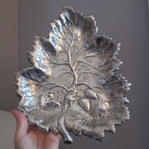 May include: A silver leaf-shaped serving dish with a raised design of a tree branch and leaves. There are three small strawberries on the branch.