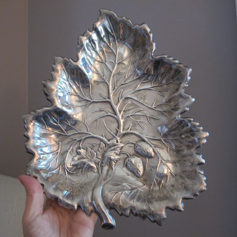 May include: A silver leaf-shaped serving dish with a raised design of a tree branch and leaves. There are three small strawberries on the branch.