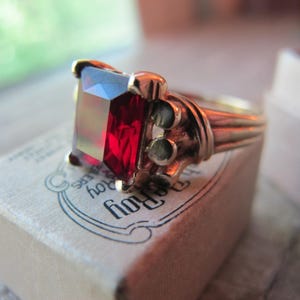 Może przedstawiać: Złoty pierścionek z dużym, prostokątnym, czerwonym kamieniem szlachetnym. Pierścionek jest osadzony w prostym, eleganckim wzorze.