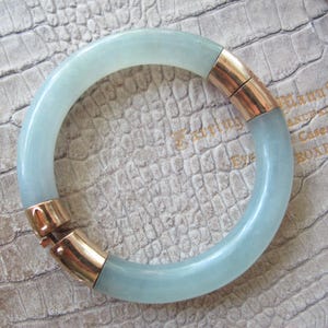 May include: A light blue jade bangle bracelet with gold tone clasps. The bracelet is on a white textured surface.